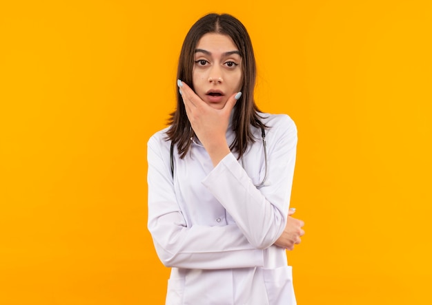 Young female doctor in white coat with stethoscope around her neck looking to the front with pensive expression puzzled standing over orange wall