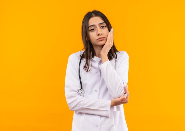 Молодая женщина-врач в белом халате со стетоскопом на шее смотрит вперед с задумчивым выражением лица, стоя над оранжевой стеной