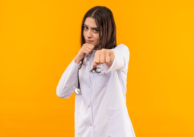 Молодая женщина-врач в белом халате со стетоскопом на шее смотрит вперед с серьезным лицом, позирующим, как боксер, стоящий над оранжевой стеной