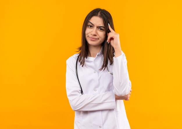 오렌지 벽 위에 서있는 회의적인 표정으로 앞으로 찾고 그녀의 목에 청진기와 흰색 코트에 젊은 여성 의사