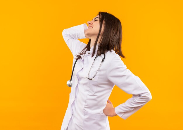 Young female doctor in white coat with stethoscope around her neck looking unwell touching her back feeling pain standing over orange wall