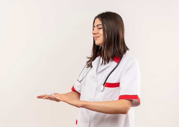 Young female doctor in white coat with stethoscope around her neck making calm down gesture with hands standing over white wall