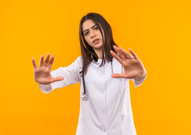 Молодая женщина-врач в белом халате со стетоскопом на шее делает защитный жест руками, смотрящими вперед, с серьезным лицом, стоящим над оранжевой стеной