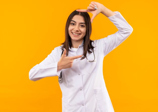 Young female doctor in white coat with stethoscope around her neck making frame with fingers looking to the front through this frame smiling standing over orange wall