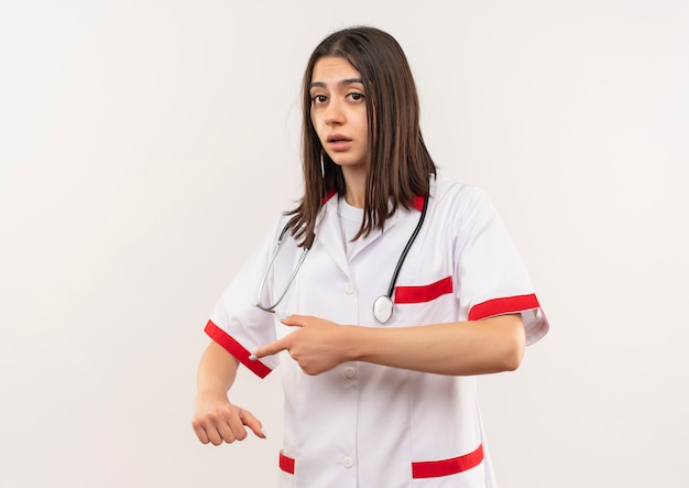 Young female doctor in white coat with stethoscope around her neck pointing to her arm reminding about time looking worried standing over white wall