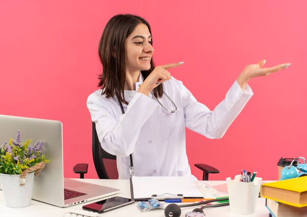 Young female doctor in white coat with stethoscope around her neck pointing with finger to the side presenting with arm of her hand smiling sitting at the table with laptop over pink wall