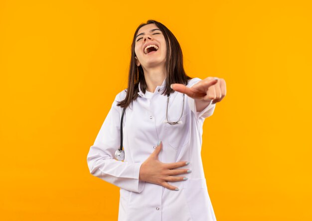 オレンジ色の壁の上に立って笑っているあなたに指で指して彼女の首の周りに聴診器を持つ白衣の若い女性医師