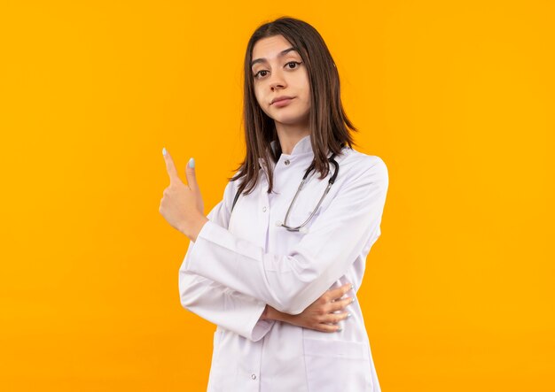 Young female doctor in white coat with stethoscope around her neck pointing with index finger to the side looking confident standing over orange wall