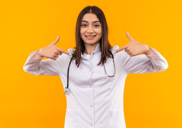 오렌지 벽 위에 서있는 자신을 검지 손가락으로 가리키는 그녀의 목에 청진기와 흰색 코트에 젊은 여성 의사