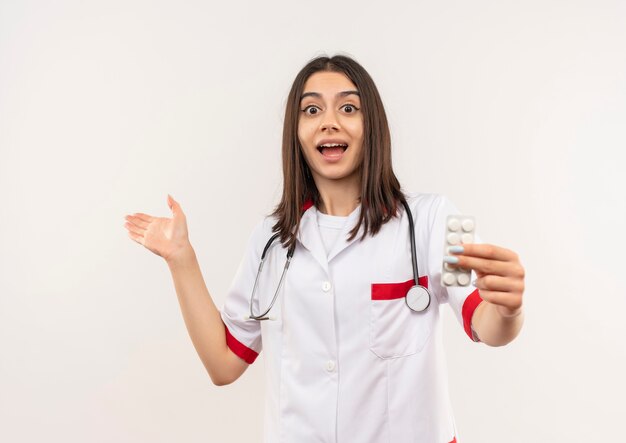 Young female doctor in white coat with stethoscope around her neck showing blister with pills pointing with arm of hand to the side happy and surprised standing over white wall