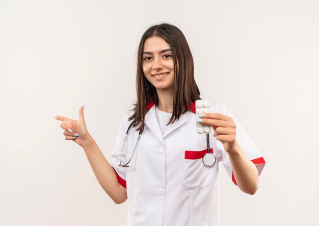 Молодая женщина-врач в белом халате со стетоскопом на шее показывает волдырь с таблетками, указывая пальцем в сторону, улыбаясь, стоя над белой стеной