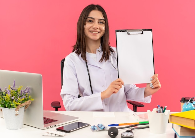 Young female doctor in white coat with stethoscope around her neck showing clipboard with blank pages smiling looking to the front sitting at the table with laptop over pink wall