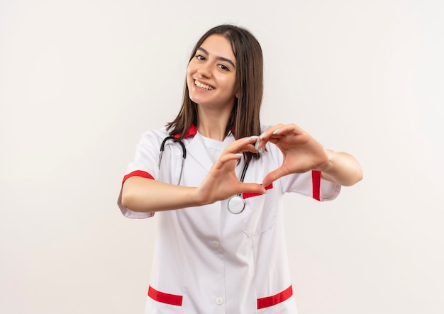 Young female doctor in white coat with stethoscope around her neck showing heart gesture smiling friendly standing over white wall