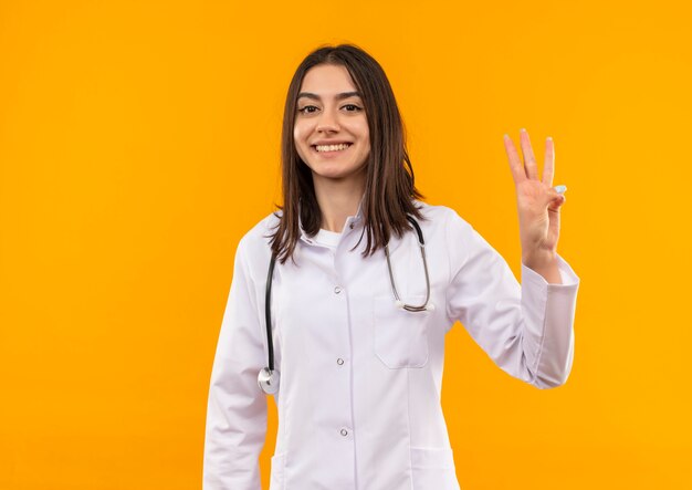 Молодая женщина-врач в белом халате со стетоскопом на шее показывает и показывает пальцами номер три, улыбаясь со счастливым лицом, стоящим над оранжевой стеной