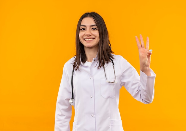 그녀의 목에 청진기와 흰색 코트에 젊은 여성 의사 표시 및 오렌지 벽 위에 서 행복 한 얼굴로 웃 고 3 번 손가락으로 가리키는