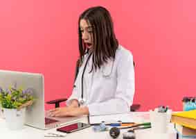 Foto gratuita giovane medico femminile in camice bianco con lo stetoscopio intorno al collo seduto al tavolo con il computer portatile che sembra confuso e sorpreso sul muro rosa