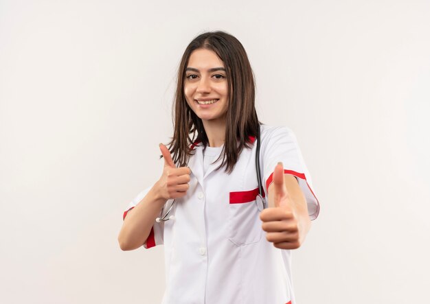 Young female doctor in white coat with stethoscope around her neck smiling cheerfully showing thumbs up standing over white wall