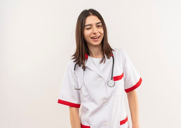 Young female doctor in white coat with stethoscope around her neck smiling and winking looking to the front standing over white wall