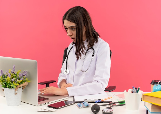 Молодая женщина-врач в белом халате со стетоскопом на шее работает на портативном компьютере, выглядит смущенным, сидя за столом над розовой стеной