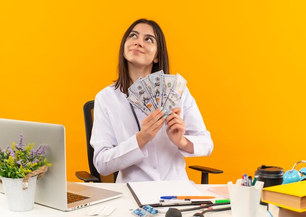 Young female doctor in white coat with stethoscope holding cash with dreamy look, sitting at the table with laptop and documents over orange wall