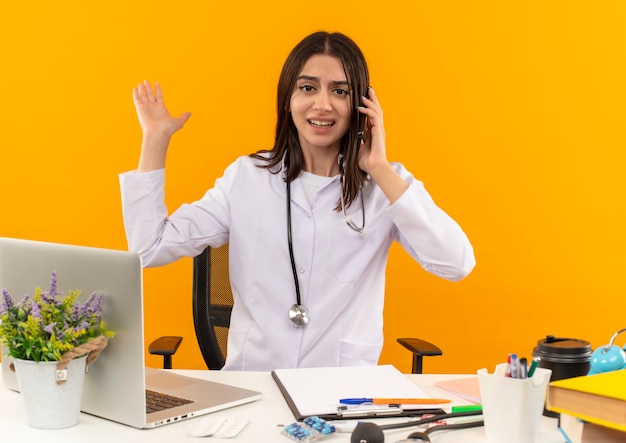 Free photo young female doctor in white coat with stethoscope talking on mobile phone looking confused sitting at the table with laptop and documents over orange wall