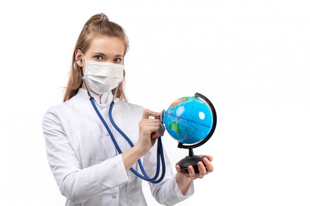 young female doctor in white medical suit in white protective mask listening to the little globe through stethoscope on the white