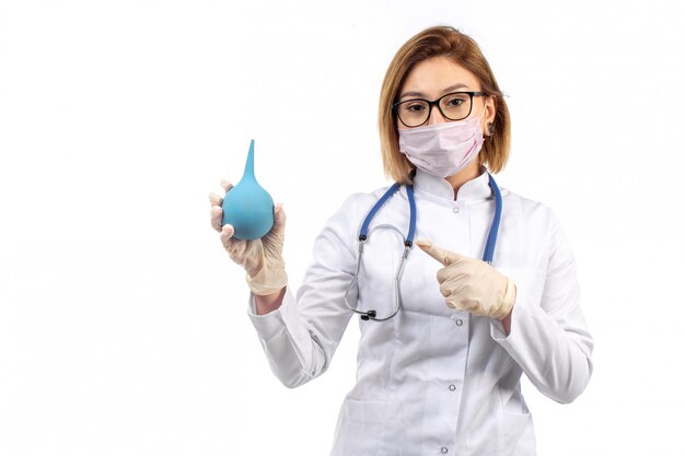 young female doctor in white medical suit with stethoscope in white protective mask holding enema on the white