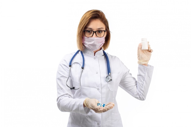 Молодая женщина-врач в белом медицинском костюме со стетоскопом в белой защитной маске держит таблетки на белом