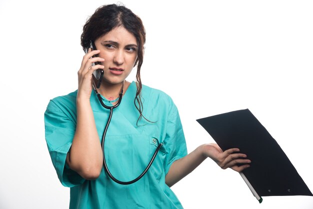 Young female doctor with clipboard and talking on cell phone on white background