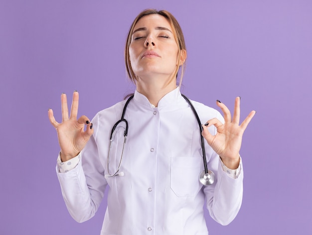 Young female doctor with closed eyes wearing medical robe with stethoscope showing meditation gesture isolated on purple wall