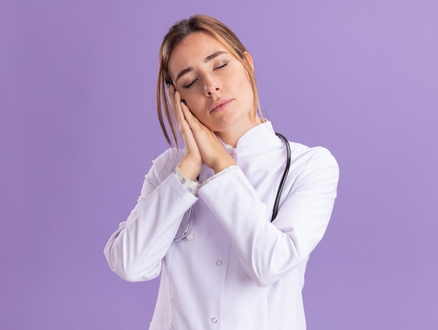 Young female doctor with closed eyes wearing medical robe with stethoscope showing sleep gesture isolated on purple wall