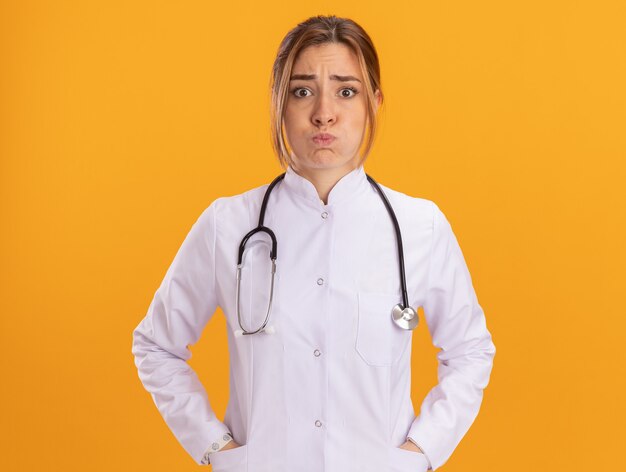 Молодая женщина-врач с пухлыми щеками в медицинском халате со стетоскопом, положив руки в карман, изолированный на желтой стене