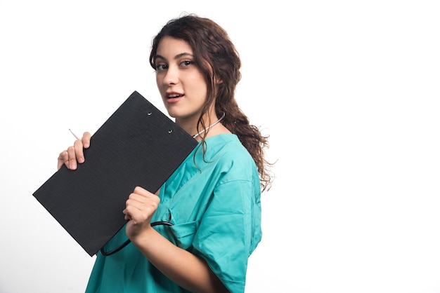 Giovane medico femminile con lo stetoscopio che tiene appunti in uniforme su priorità bassa bianca