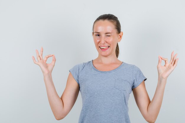 Young female doing meditation and winking eye in grey t-shirt front view.