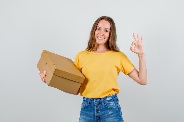 Young female doing ok sign with cardboard box in t-shirt, shorts and looking glad. front view.