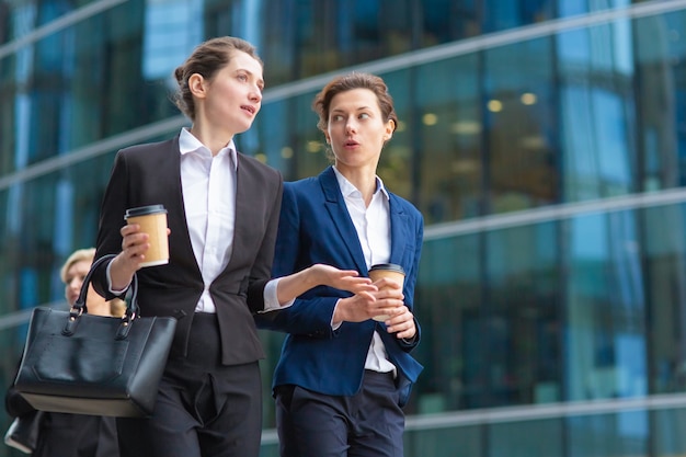 Young female employees with paper coffee cups wearing office suits, walking together past glass office building, talking, discussing project. Low angle. Work break or friendship concept
