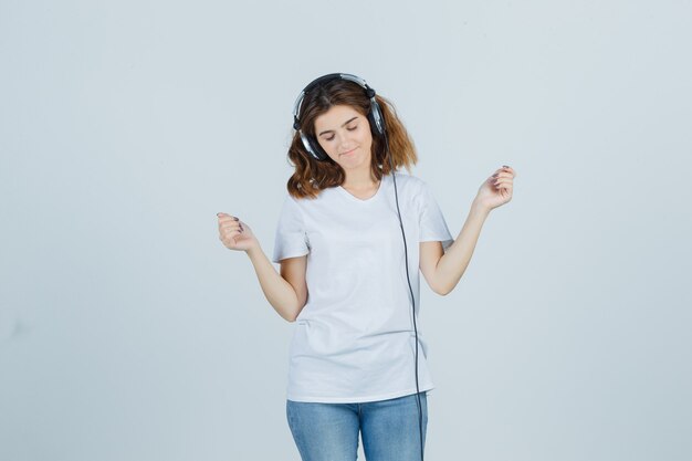 白いTシャツ、ジーンズ、陽気に見えるヘッドフォンで音楽を楽しんでいる若い女性。正面図。