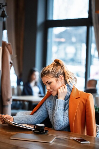 Young female entrepreneur relaxing on coffee break and reading daily newspaper in cafeteria