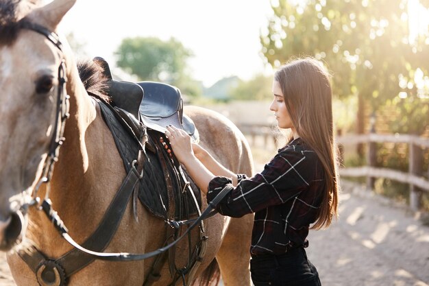 Young female equine owner tying down the saddle preparing to take a ride down to the river from the animal farm on a sunny autumn day