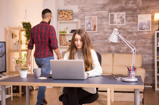 Young female freelancer working on laptop in home office. Boyfriend in the background.