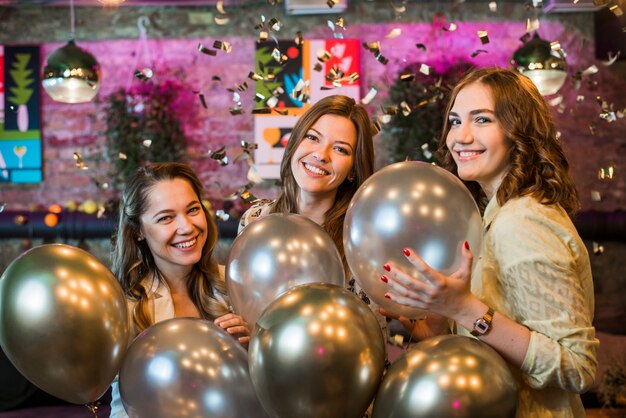 Молодые подруги держа серебряные воздушные шары наслаждаясь в партии