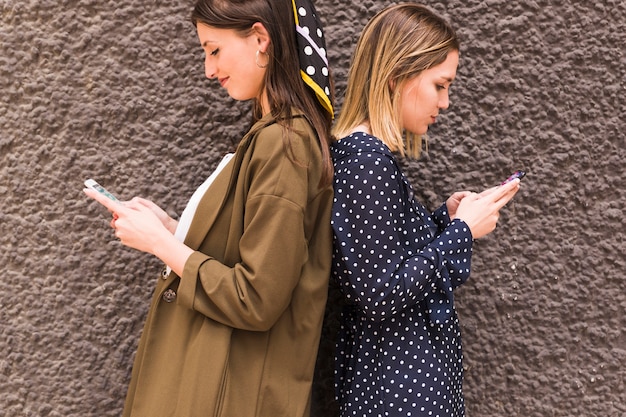 young female friends standing back to back using mobile phone