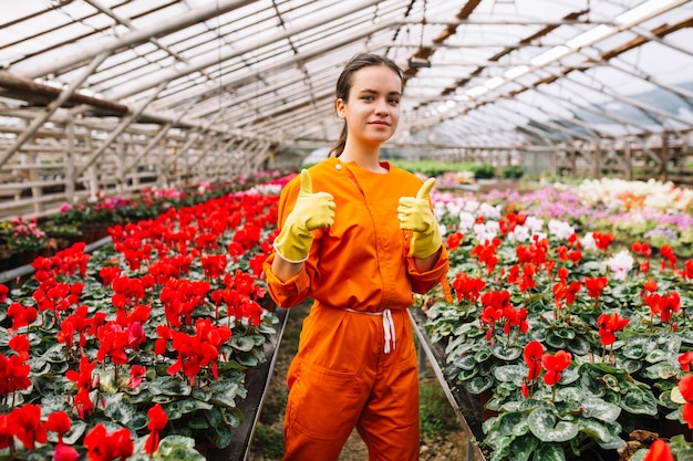 Young female gardener gesturing thumbs up with fresh flowers growing in greenhouse