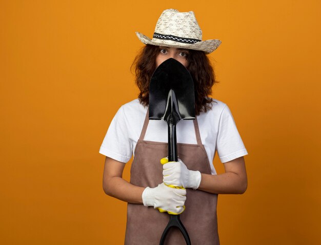 young female gardener in uniform wearing gardening hat and gloves covered face with spade