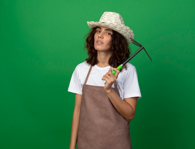 young female gardener in uniform wearing gardening hat holding hoe rake