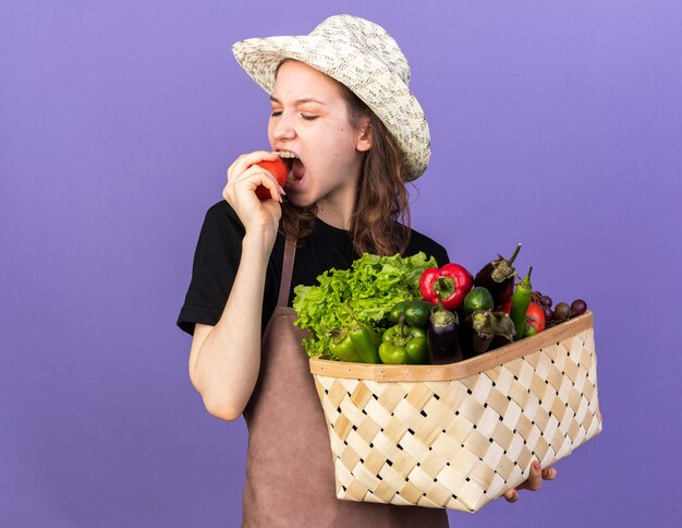 Молодая женщина-садовник в садовой шляпе держит корзину с овощами и кусочки помидора