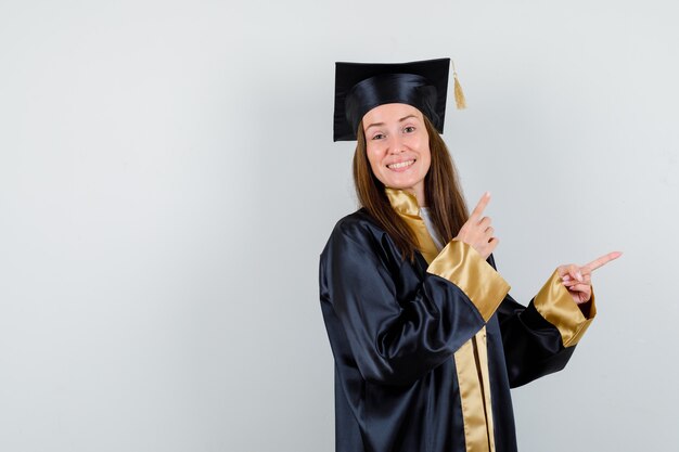 Young female graduate in academic dress pointing aside and looking pretty , front view.