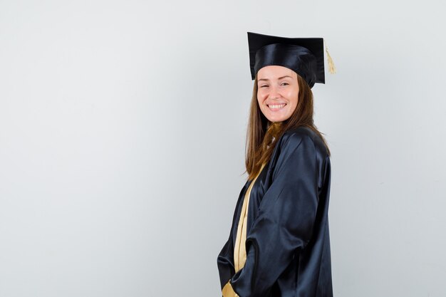 Young female graduate in academic dress posing while standing and looking happy , front view.