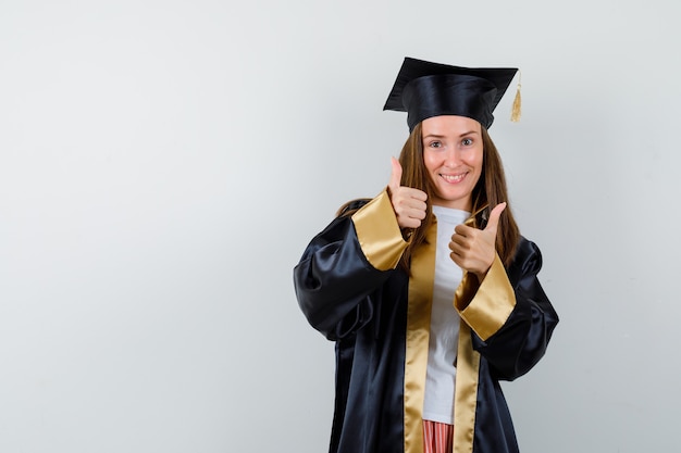 Young female graduate in academic dress showing thumps up and looking blissful , front view.