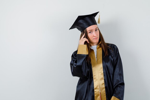 Young female graduate holding finger on temple in academic dress and looking pensive. front view.